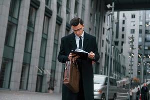 With notepad in hands. Businessman in black suit and tie is outdoors in the city photo