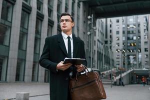 With notepad in hands. Businessman in black suit and tie is outdoors in the city photo