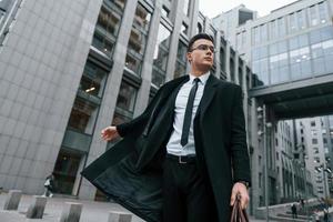 edificio gris detrás. hombre de negocios con traje negro y corbata está al aire libre en la ciudad foto