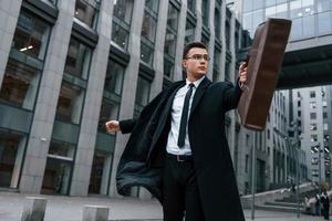 Grey building behind. Businessman in black suit and tie is outdoors in the city photo