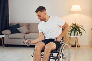 Doing exercises by using dumbbells. Disabled man in wheelchair is at home photo