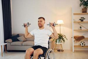 Doing exercises by using dumbbells. Disabled man in wheelchair is at home photo