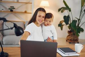 sentado junto a la mesa con la computadora portátil. madre con su pequeña hija está en casa juntos foto