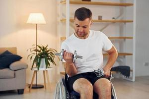 Lifting dumbbells. Disabled man in wheelchair is at home photo