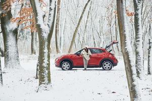 Rural scene. Beautiful young woman is outdoors near her red automobile at winter time photo