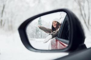 View in the side mirror. Beautiful young woman is outdoors near her red automobile at winter time photo