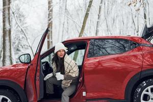 New car is in the forest. Beautiful young woman is outdoors near her red automobile at winter time photo
