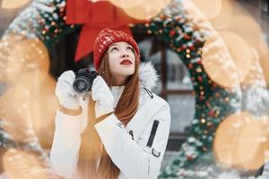 With camera in hands. Happy young woman standing outdoors and celebrating christmas holidays photo