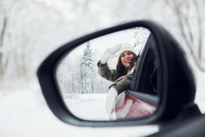 View in the side mirror. Beautiful young woman is outdoors near her red automobile at winter time photo