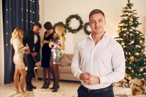 Man in formal clothes standing in front of his friends. Group of people have a new year party indoors together photo