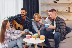 Man is playing acoustic guitar. Group of friends have party indoors together photo