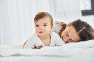 On the white bed. Mother with her little daughter is indoors at home together photo