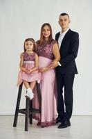 Girl is sitting on the chair. Happy couple with little daughter is standing in the studio photo