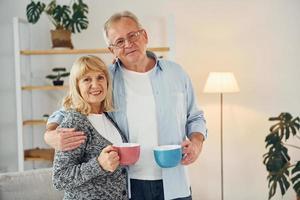 Standing and holding cups of drinks. Senior man and woman is together at home photo