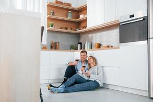 Drinking wine. Couple sitting on the floor of modern kitchen photo