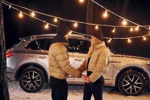 With modern car behind. Couple standing in the forest and celebrating New year photo