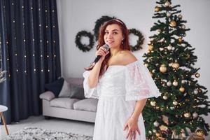 Standing in the christmas decorated domestic room. Woman in white dress and with microphone in hands is singing in the photo