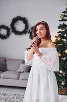 Standing in the christmas decorated domestic room. Woman in white dress and with microphone in hands is singing in the photo