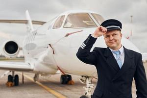 Posing for a camera. Experienced pilot in uniform standing outside near plane photo