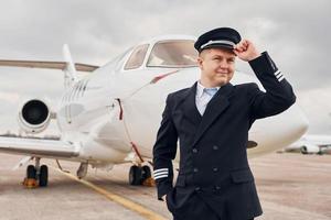 Posing for a camera. Experienced pilot in uniform standing outside near plane photo