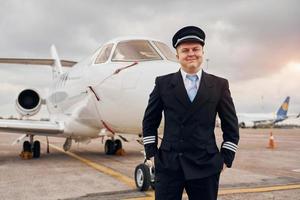 posando para una cámara. piloto experimentado en uniforme parado afuera cerca del avión foto