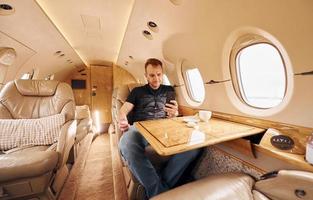 Passenger in casual clothes sits by table with phone in hands. In airplane photo