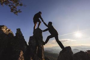 silueta de dos personas escalando el acantilado de la montaña y una de ellas dando la mano. gente ayudando y, concepto de trabajo en equipo. foto