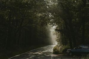 Blue car parked on the edge of the forest by the road with sun rays of morning sun shining through the trees in the background photo