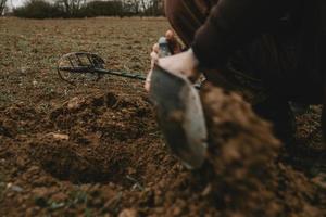 Detail of man's hands with shovel in it, during digging a hole in ground after positive signal of metal detector with lying metal detector in background photo