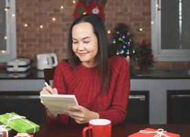 mujer asiática con suéter de punto rojo sentada en la mesa con una taza roja de café y cajas de regalo en la cocina con decoración navideña, usando una lista de nombres para escribir en un cuaderno. foto