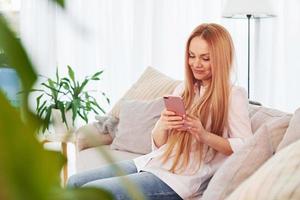 With phone in hands. Young woman in white shirt and jeans is at home photo