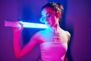 In glasses. Fashionable young woman standing in the studio with neon light photo