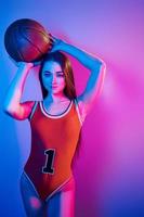 In basketball uniform. Fashionable young woman standing in the studio with neon light photo