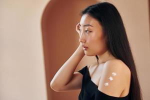 Uses beauty cream. Young serious asian woman standing indoors photo