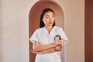 Young doctor. Serious asian woman standing indoors photo