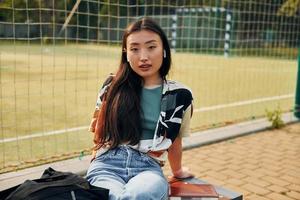 Sits against sport field. Young asian woman is outdoors at daytime photo