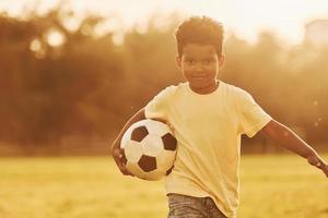 joven futbolista. niño afroamericano se divierte en el campo durante el día de verano foto