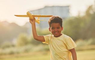 Yellow plane in hands. African american kid have fun in the field at summer daytime photo