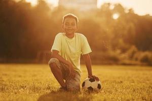 se sienta con balón de fútbol. niño afroamericano se divierte en el campo durante el día de verano foto