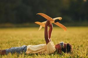 tumbado con un avión de juguete en el césped. niño afroamericano se divierte en el campo durante el día de verano foto