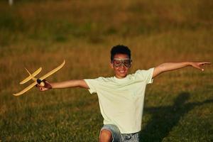 In casual clothes with toy plane. African american kid have fun in the field at summer daytime photo