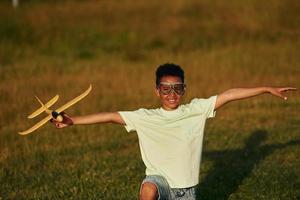 In casual clothes with toy plane. African american kid have fun in the field at summer daytime photo