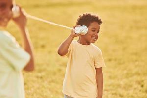 tener una conversación usando tazas en los nudos. dos niños afroamericanos se divierten juntos en el campo durante el día de verano foto