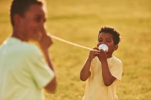 tener una conversación usando tazas en los nudos. dos niños afroamericanos se divierten juntos en el campo durante el día de verano foto