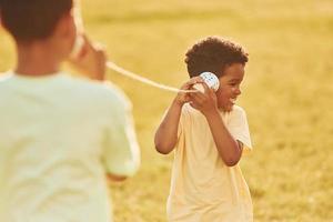 tener una conversación usando tazas en los nudos. dos niños afroamericanos se divierten juntos en el campo durante el día de verano foto