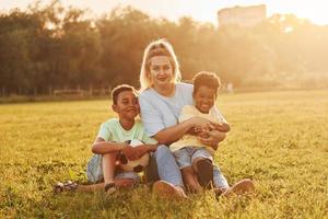 Adult woman sits with two black kids on the field at summer daytime photo