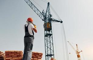 With crane at background. Young construction worker in uniform is busy at the unfinished building photo