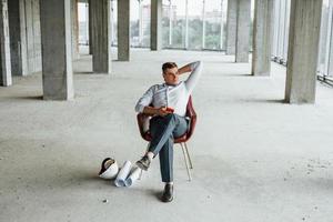 Sits on the chair. Young man in formal wear is working indoors on the construction photo