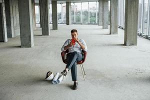 Sits on the chair. Young man in formal wear is working indoors on the construction photo