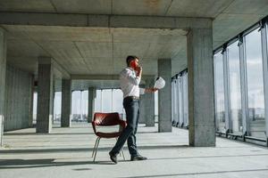 tener una conversación usando el teléfono. un joven con ropa formal está trabajando en el interior de la construcción foto